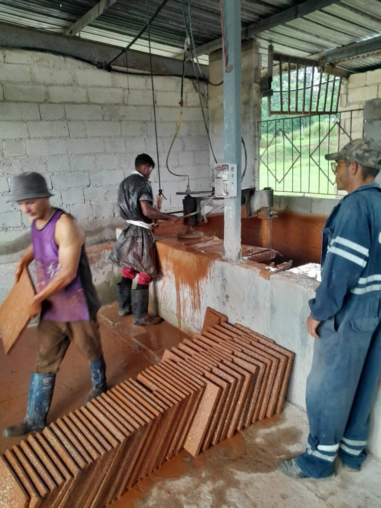 La imagen muestra un taller artesanal con paredes de bloques y techo de metal, donde tres hombres trabajan con barro. Dos de ellos moldean y trasladan piezas rectangulares de arcilla húmeda, mientras un tercero observa el proceso. En el suelo hay filas de piezas recién formadas, de color rojizo, y el piso está mojado con barro, lo que refleja un trabajo manual intenso en un entorno rural visible a través de una ventana con rejas.