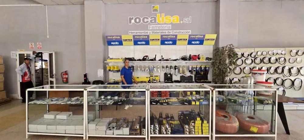 Fotografía del interior de una ferretería amplia y bien iluminada. En primer plano se observan vitrinas de vidrio con herramientas, accesorios eléctricos y materiales de construcción organizados. Detrás del mostrador hay una persona atendiendo. En la pared del fondo se leen los rótulos “Rocalisa S.R.L.” y “Ferretería”, junto a estanterías con herramientas, cables y equipos industriales.