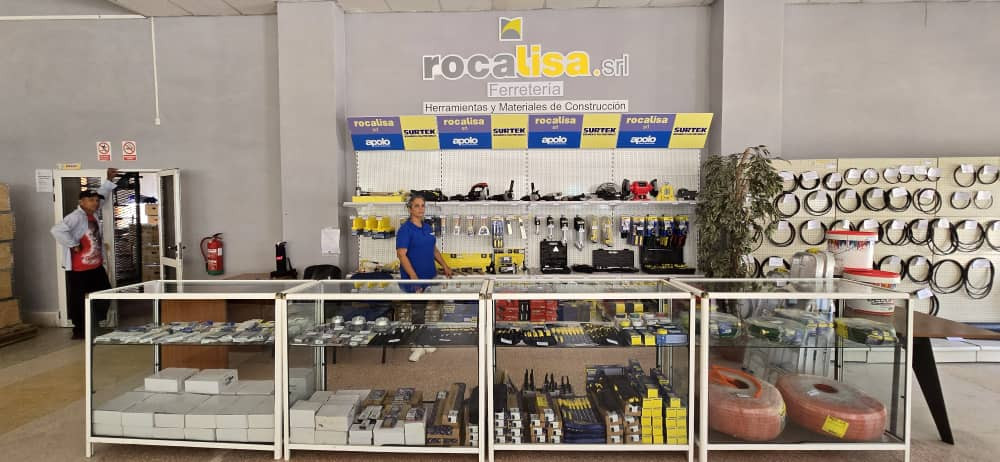 Fotografía del interior de una ferretería amplia y bien iluminada. En primer plano se observan vitrinas de vidrio con herramientas, accesorios eléctricos y materiales de construcción organizados. Detrás del mostrador hay una persona atendiendo. En la pared del fondo se leen los rótulos “Rocalisa S.R.L.” y “Ferretería”, junto a estanterías con herramientas, cables y equipos industriales.
