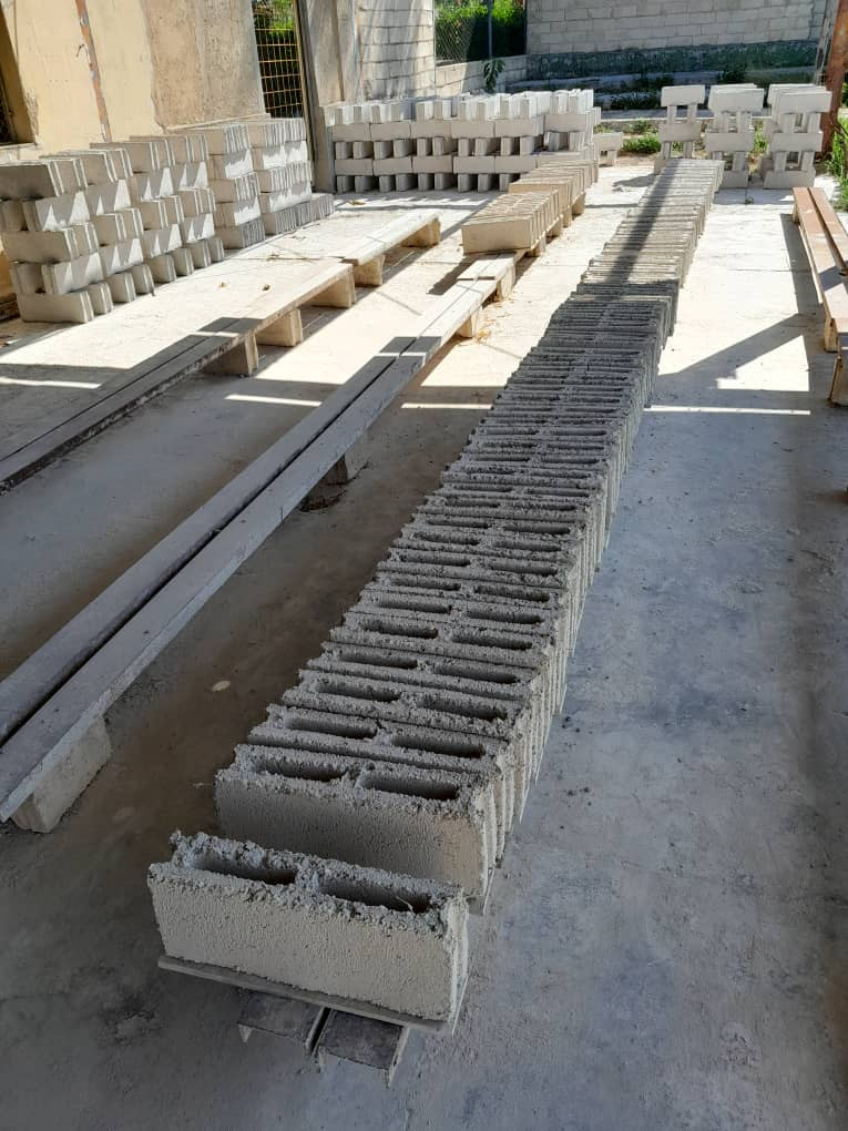 Fotografía de un área de trabajo al aire libre donde se observan largas filas de bloques prefabricados de hormigón con cavidades internas, colocados sobre el suelo para su secado. En el fondo se aprecian más bloques apilados, tablas de madera y una estructura de paredes sin terminar, lo que sugiere un entorno de producción o almacenamiento de materiales de construcción.