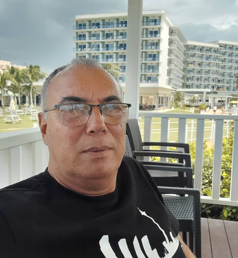 Fotografía tipo autorretrato de un hombre adulto (Rolando Rodríguez) sentado en una terraza al aire libre. Lleva gafas y una camiseta oscura con un diseño gráfico claro. Al fondo se observan edificios modernos de varios pisos, áreas verdes y mobiliario exterior, lo que sugiere un entorno hotelero o turístico. La imagen transmite un ambiente relajado y cotidiano.