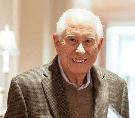 La imagen muestra a Carmelo Mesa-Lago, en un espacio interior con iluminación cálida. Aparece sonriente, con cabello blanco, vestido con un saco de cuadros en tonos tierra sobre un suéter marrón y camisa clara, lo que proyecta una imagen formal y sobria. El fondo está desenfocado.