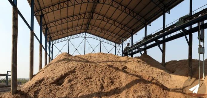 Bajo una gran estructura metálica con techo alto, se observan enormes montículos de bagazo de color beige. A la derecha hay una cinta transportadora industrial elevada. El lugar parece una planta o almacén al aire libre destinado al procesamiento o almacenamiento de bagazo.