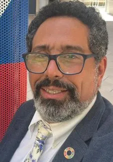 Retrato de primer plano de un hombre maduro de piel morena, cabello rizado canoso y barba frondosa recortada también con canas. Lleva espejuelos de armazón oscuro, una camisa blanca con corbata de estampado floral en tonos azules y una chaqueta de vestir gris. En la solapa de la chaqueta lleva un pin circular multicolor que representa los Objetivos de Desarrollo Sostenible (ODS).
