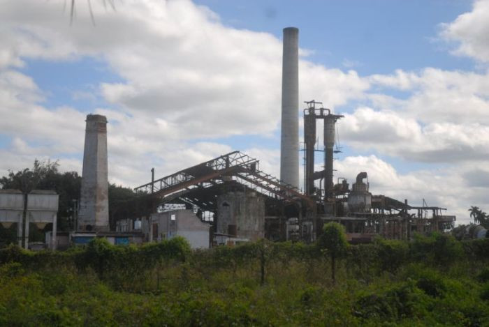 The image shows an abandoned industrial plant in ruins, with rusted structures and vegetation growing around it. It evokes a sense of neglect and the end of a once-productive era.