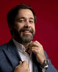 A half-body portrait of a man with a beard and brown hair, smiling at the camera. He is wearing a white shirt, open at the neck, and a textured blue jacket. With his right hand, he adjusts the lapel of his jacket, and with his left, the collar of his shirt, revealing a silver wristwatch. The background is a solid, vibrant red.