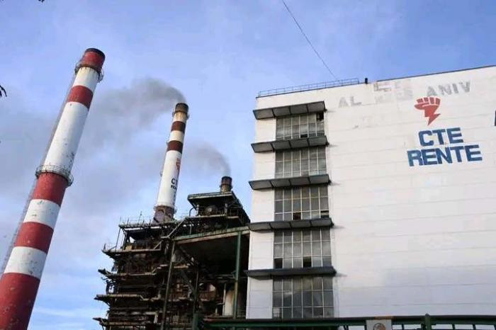 Fotografía de una central termoeléctrica con dos altas chimeneas cilíndricas que emiten humo oscuro. A un lado se observa un edificio industrial de varios pisos con el logotipo de la Central de Trabajadores de Cuba en la fachada. La imagen muestra instalaciones envejecidas y elementos visibles de combustión, representando la generación eléctrica basada en combustibles fósiles.