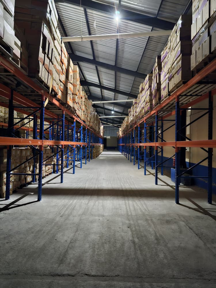 Interior de un almacén con pasillos largos y simétricos, flanqueados por estanterías metálicas de varios niveles cargadas con cajas de cartón apiladas. La estructura industrial —techo alto de láminas metálicas y vigas expuestas— está iluminada por luces cenitales que refuerzan la profundidad del espacio. El piso despejado y la organización uniforme sugieren un sistema logístico orientado al almacenamiento y distribución a escala.