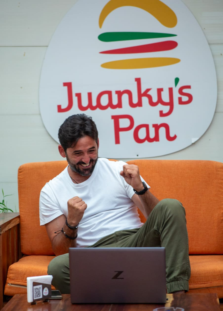 Hombre sentado en un sofá naranja usando una computadora portátil, sonriente y con gesto de celebración. Detrás se observa un letrero con el logo y el nombre ‘Juanky’s Pan’.