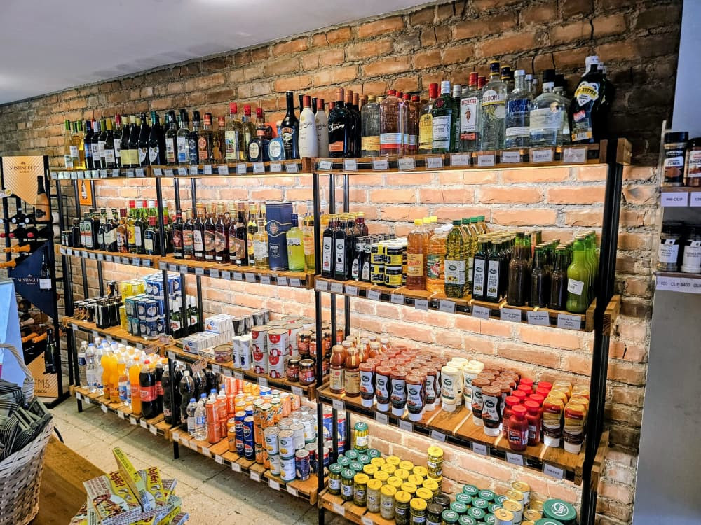 Estanterías de madera y metal apoyadas sobre una pared de ladrillo visto exhiben una amplia variedad de productos alimenticios y bebidas. En los niveles superiores predominan botellas de vinos y licores, mientras que en los inferiores se organizan aceites, salsas, conservas y refrescos, todos alineados con etiquetas visibles y precios señalizados. La iluminación uniforme resalta la abundancia y diversidad de la oferta en un entorno comercial ordenado.