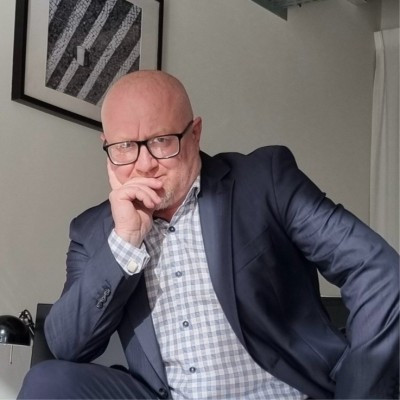 Photograph of a middle-aged man seated indoors, wearing glasses, a dark blazer, and a light-colored checkered shirt. He is bald and rests one hand against his chin in a thoughtful pose, looking directly toward the camera. The setting appears to be a modern interior, with a framed artwork on the wall behind him and soft, natural lighting, conveying a reflective and serious mood.
