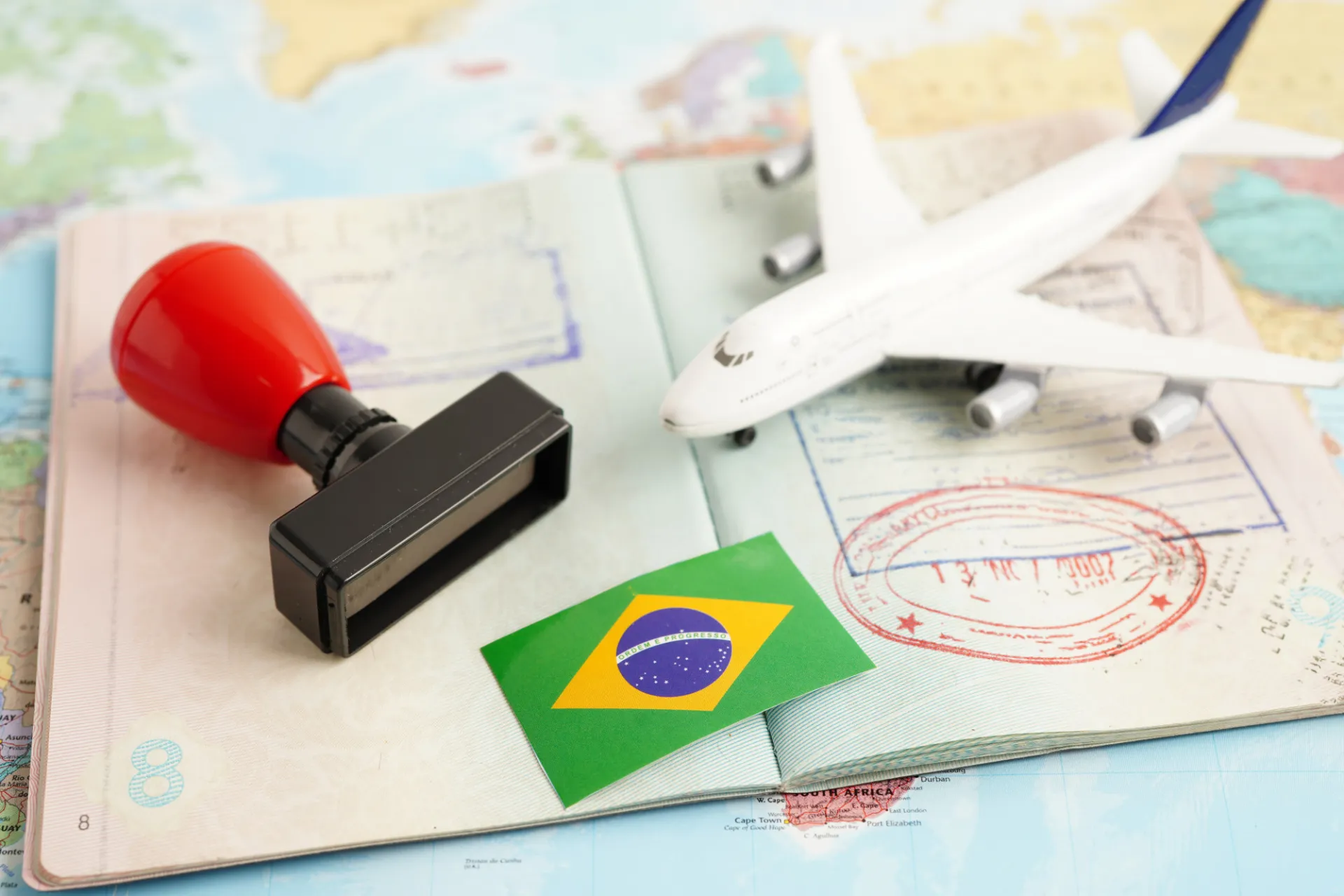 Un pasaporte abierto con sellos de entrada y salida, junto a un sello de tinta, un avión en miniatura y una pequeña bandera de Brasil sobre un mapa. La imagen representa el proceso de los viajes internacionales, así como los procedimientos relacionados con la inmigración o la migración.