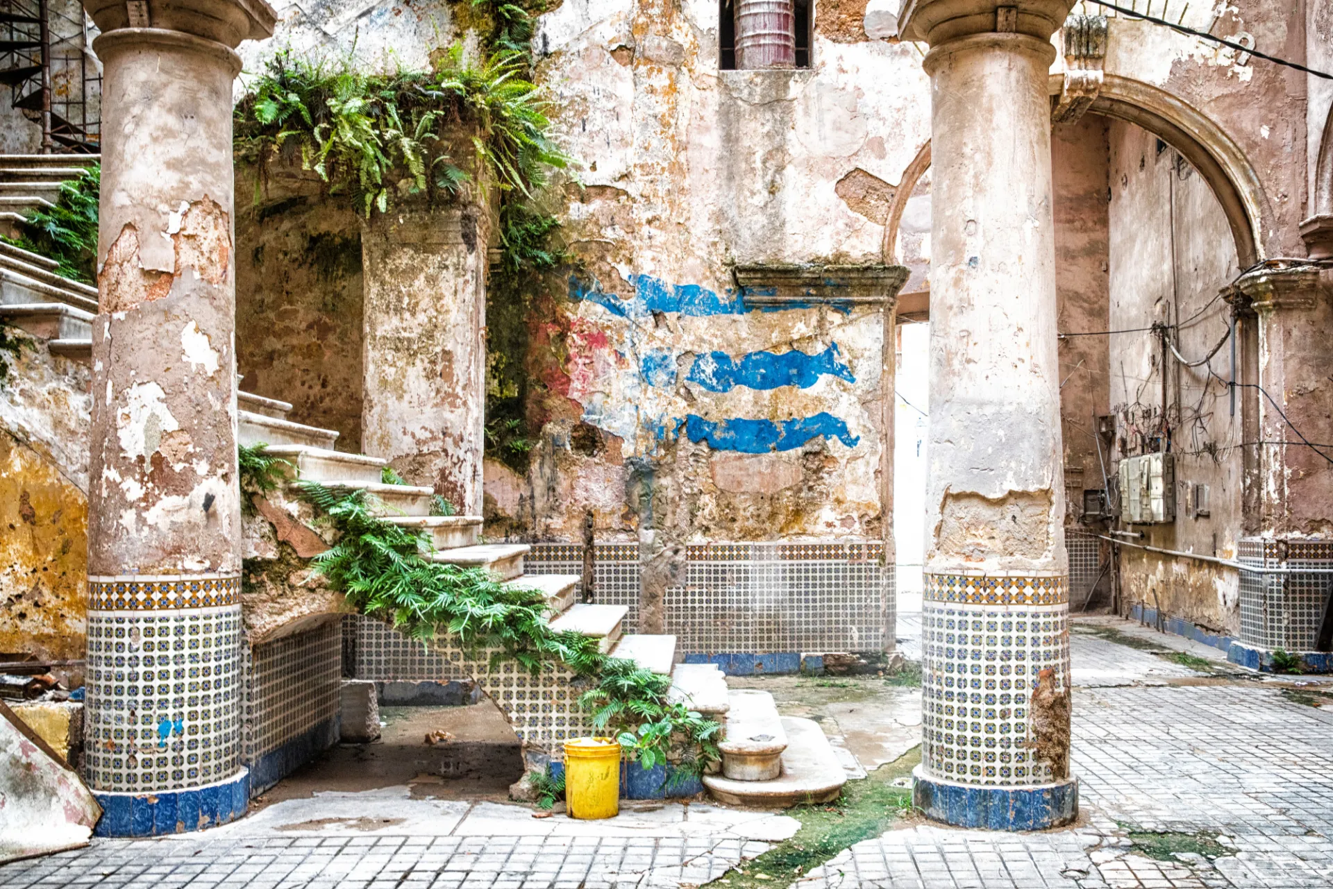 Fotografía de un patio interior deteriorado en Cuba, que muestra un edificio antiguo con columnas desgastadas decoradas con azulejos, paredes agrietadas y descascaradas, y una pequeña escalera parcialmente cubierta por plantas. En una de las paredes se observa una bandera cubana, descolorida y pintada o colgada sobre la superficie dañada. Cubetas y recipientes reposan sobre el piso de azulejos, y la escena transmite la sensación de arquitectura envejecida, abandono y el paso del tiempo.
