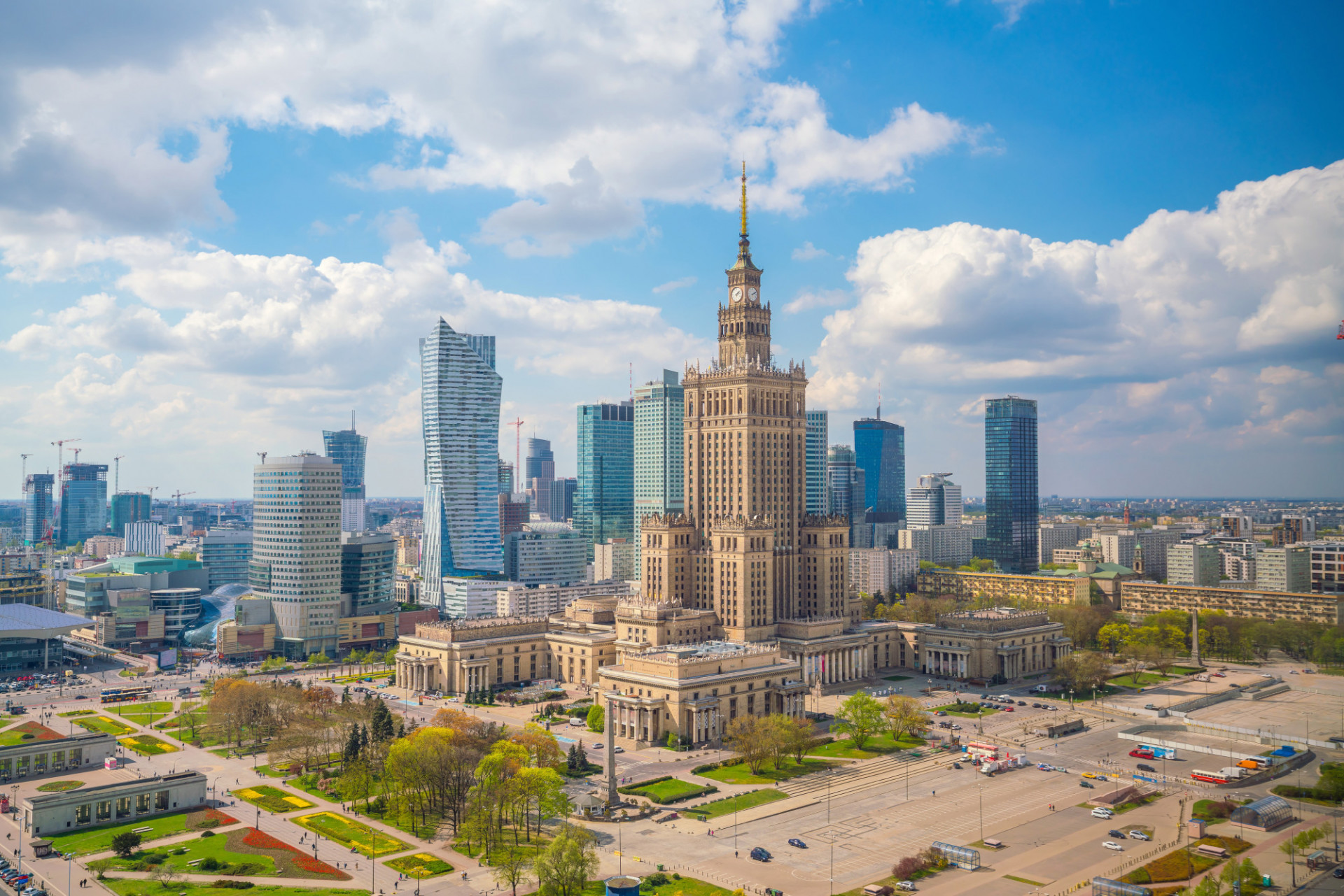 Panoramic photograph of the city of Warsaw, seen from an elevated viewpoint, with wide avenues and modern buildings extending around it. At the center stands a tall, symmetrical historic building in a light color, featuring a central tower topped with a spire, surrounded by green areas and large open plazas. Around it, contemporary glass-and-steel skyscrapers, some with curved shapes, contrast with the classical architecture of the main building. 