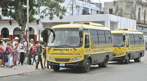 En primer plano aparecen dos microbuses amarillos identificados como cooperativas de transporte, estacionados junto a la acera. Varias personas hacen fila o suben al vehículo delantero, mientras otras esperan en la acera. Al fondo se observan edificaciones de estilo urbano, algunas deterioradas, y árboles que proyectan sombra sobre la escena. La imagen refleja un momento cotidiano del transporte público alternativo en la ciudad.