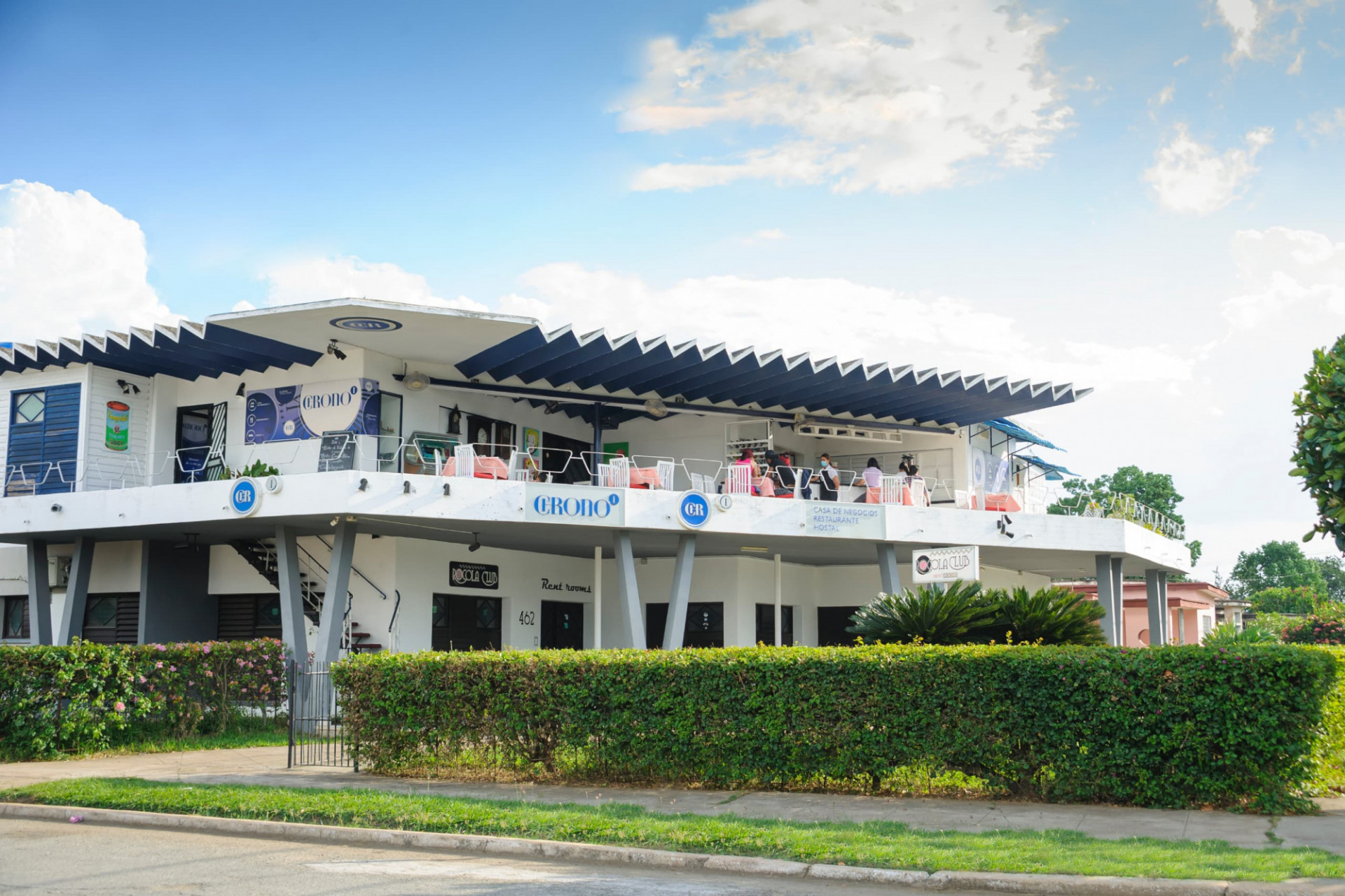 Restaurante Chrono, Camagüey edificio azul y blanco con seto verde bajo un cielo azuzl con nueves blancas