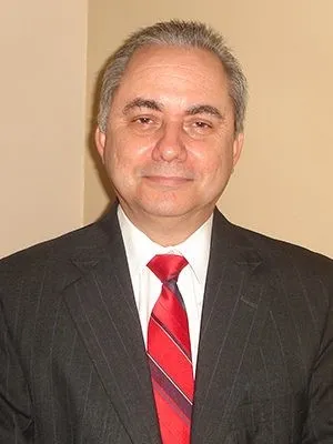 La fotografía muestra a un hombre adulto de cabello corto y canoso, vestido formalmente con un traje oscuro, camisa blanca y corbata roja con franjas diagonales. Está retratado de frente, desde el pecho hacia arriba, y mira directamente hacia la cámara con una expresión tranquila y ligeramente sonriente. El fondo es de color beige y uniforme, sin otros elementos visibles.