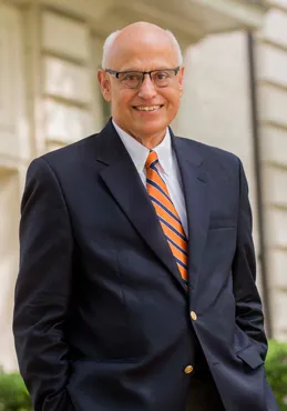 man wearing glasses, dressed in a dark suit and red tie, smiling at the camera