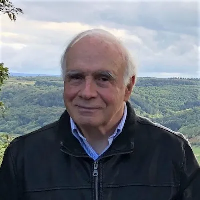 man with grey hair, smiling, wearing a dark jacket and standing before a mountainous horizon