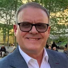 Smiling man wearing dark glasses, with a white shirt and blue jacket with trees in the background