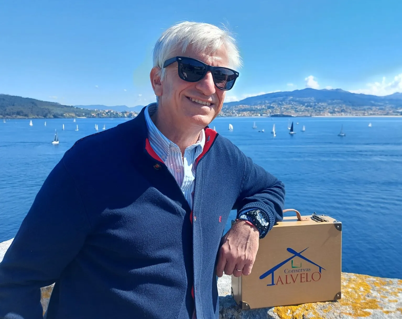 hombre de pelo blanco portando lentes oscuros frente una bahía con barcos de vela y cielo azul