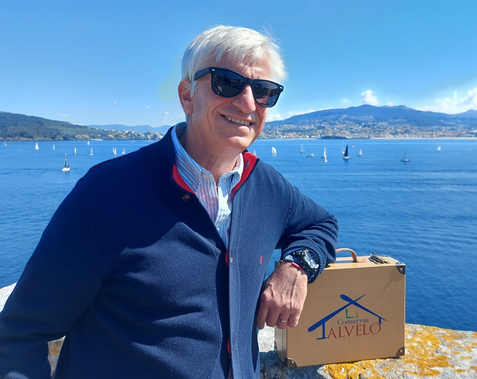 hombre de pelo blanco portando lentes oscuros frente una bahía con barcos de vela y cielo azul