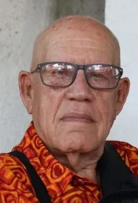 A close-up portrait of an older man looking at the camera. He is almost completely bald and wears dark-framed glasses. He is dressed in a shirt featuring a pattern of curved shapes in red, orange, and yellow. His face fills much of the frame and bears a serious, calm expression. The background is light and neutral, with no prominent features.