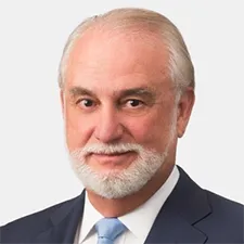 Hombre con cabello gris y barba en traje oscuro con corbata azul claro