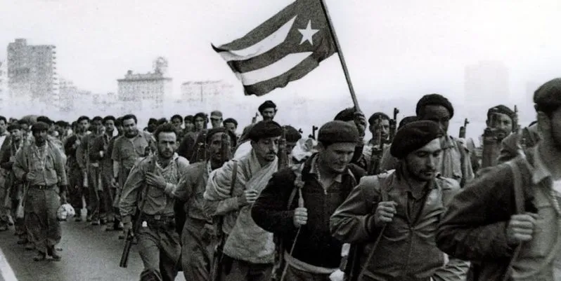 People marching along Havana’s Malecón, some carrying berets and rifles, and one carrying the Cuban flag.