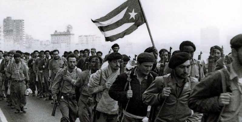 People marching along Havana’s Malecón, some carrying berets and rifles, and one carrying the Cuban flag.