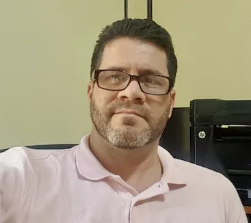hombre con barba y lentes y pelo oscuro portando camisa rosada con impresora atrás