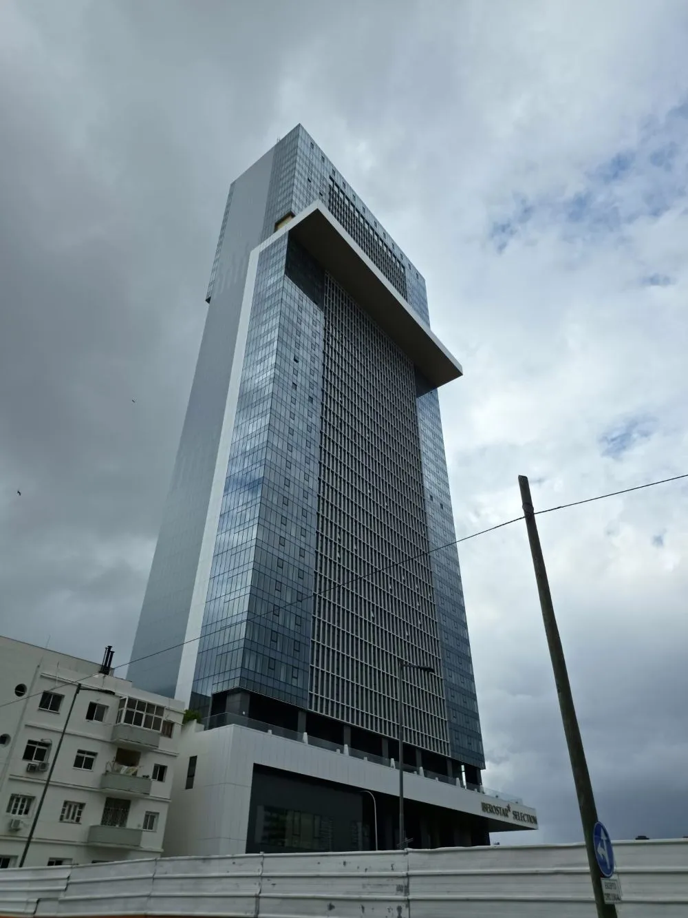 Low-angle view of the Torre K in Havana. A modern glass skyscraper with clean, straight lines and a reflective façade rises imposingly toward a cloudy sky. At the top, a horizontal structure juts out, breaking the building’s verticality. At the base, lower structures are visible along with a fence in the foreground, while a pole and cables partially cross the scene, emphasizing the urban perspective.