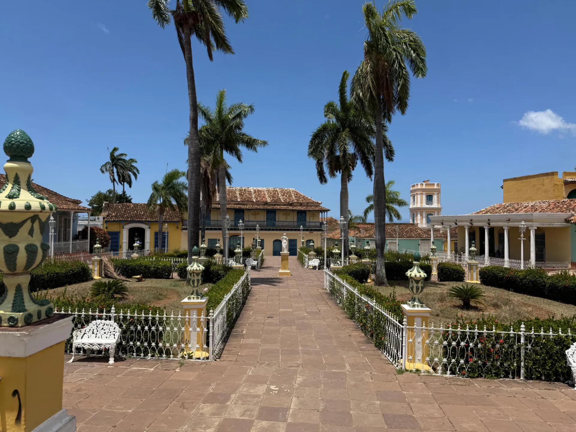 Fotografía de una plaza del centro histórico de Trinidad, Cuba, con arquitectura colonial, edificios de fachadas amarillas, techos de tejas y una torre al fondo. La plaza está prácticamente desierta de turistas, aunque se trata de un espacio que normalmente presenta una intensa afluencia de visitantes. El área ajardinada, las altas palmeras, los bancos y las rejas ornamentales contrastan con la ausencia de personas en la escena.