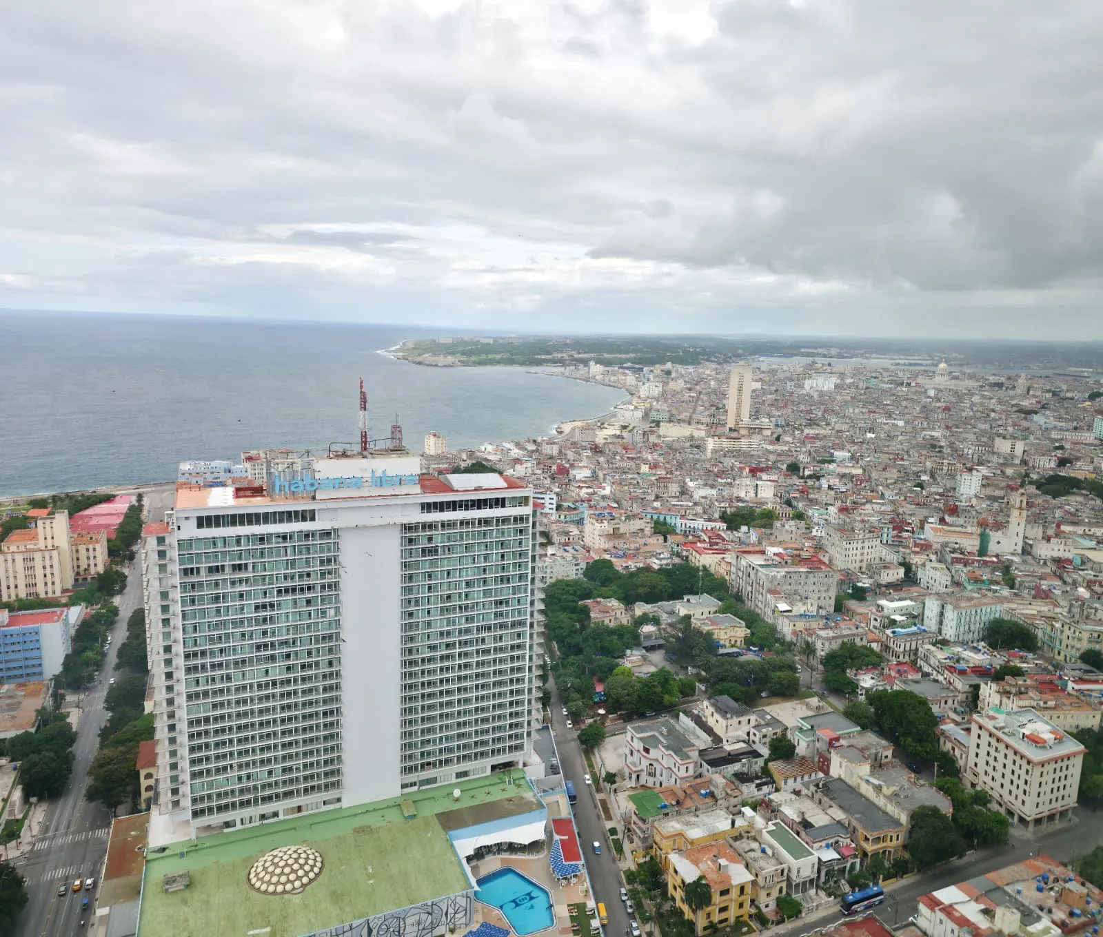 Elevated panoramic view of Havana on a cloudy day. In the foreground stands a tall building (the Habana Libre Hotel) with the sign “Habana Libre” on its rooftop. Surrounding it is a dense urban fabric of low- and mid-rise buildings, with streets and some green areas. In the background, the city stretches along the sea, forming a broad coastline that fades into the horizon beneath a sky covered in gray clouds.