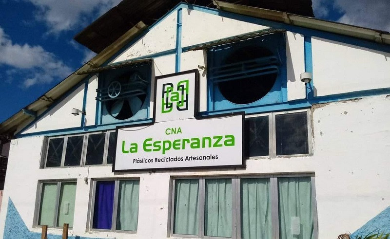 Fotografía de la fachada de una nave industrial en Cuba perteneciente a la cooperativa no agropecuaria la esperanza “La Esperanza”. El edificio es de color blanco con detalles en azul y presenta grandes ventiladores circulares en la parte superior. En el centro se observa un cartel con el nombre “CNA La Esperanza” y la frase “Plásticos Reciclados Artesanales”. Debajo, varias ventanas con cortinas de colores claros.