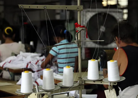 En primer plano se observan varios conos de hilo blanco colocados en una máquina industrial, con hilos tensados que se extienden hacia arriba. En el fondo, desenfocados, aparecen varios trabajadores sentados frente a máquinas de coser mientras confeccionan prendas. El ambiente sugiere un espacio de producción activa dentro de una cooperativa textil.