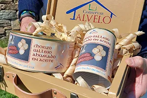Una caja con conservas aguantada por dos manos con conservas de chorizos y las palabras "conservas alvelo"
