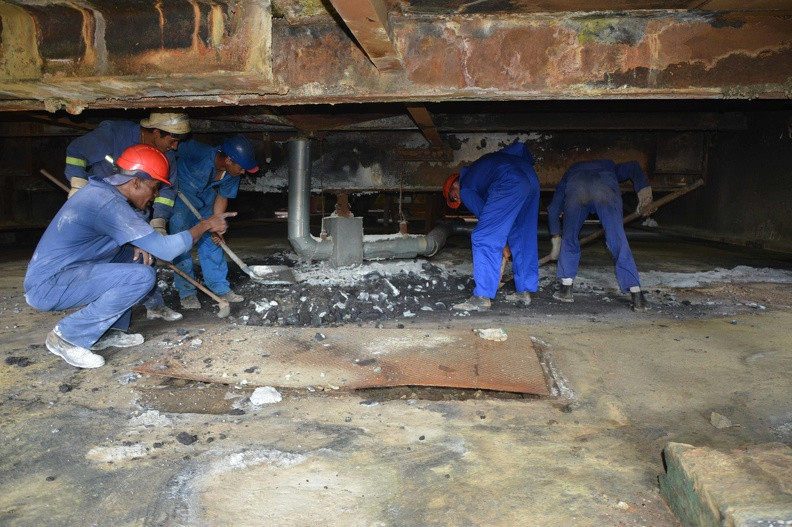 Several workers wearing helmets and blue uniforms are engaged in cleaning or repair tasks beneath a metal structure, working together in a deteriorated industrial setting.