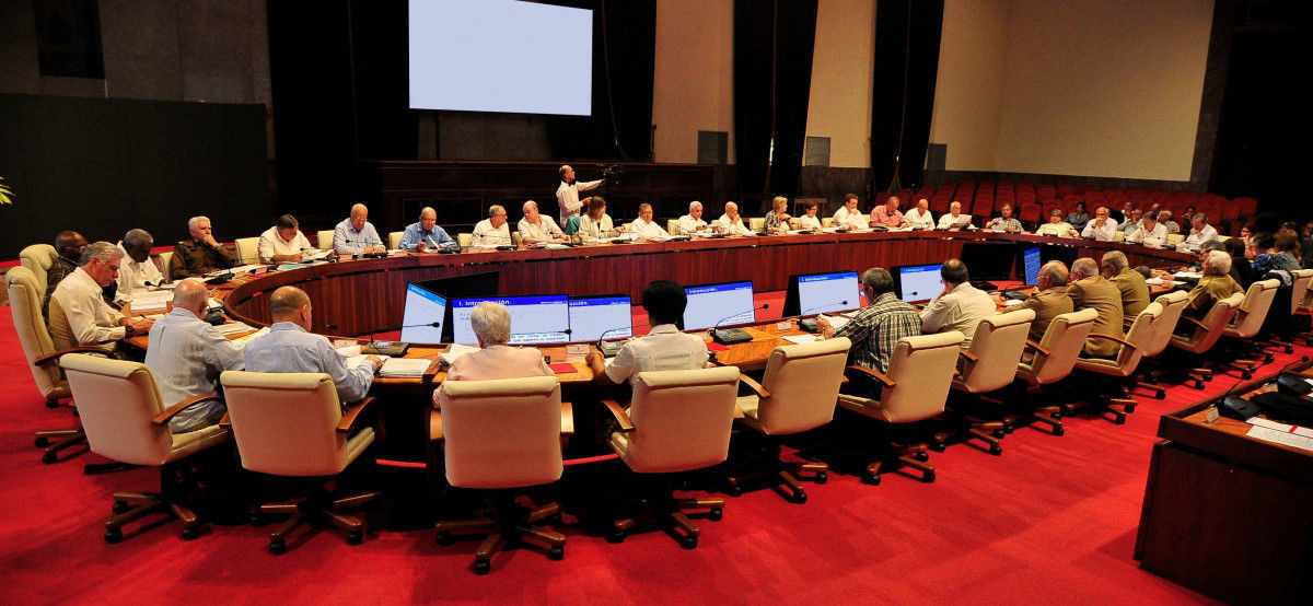 Reunión oficial con decenas de personas sentadas alrededor de una mesa ovalada, cada una con computadoras, en una sala amplia con pantalla al fondo.